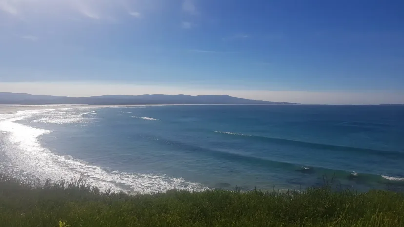 A deep blue bay curving to a distant headland, seen from a grassy clifftop.