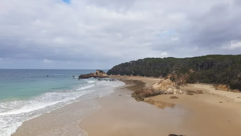 Turquoise surf breaking on sand below a rocky forested headland.